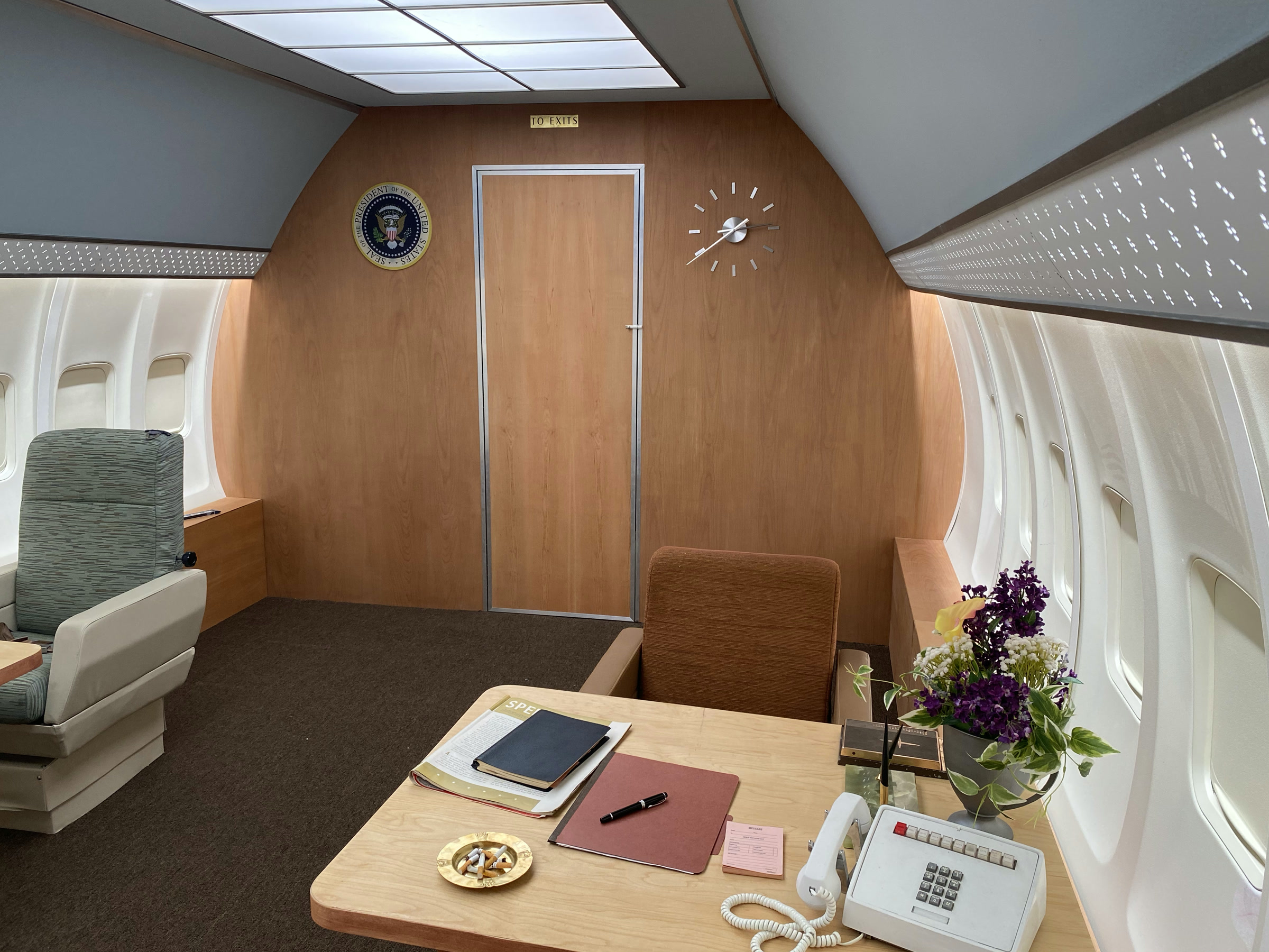 The Air Force One stateroom set: a presidential seal and sunburst clock on the wood bulkhead, a brass 'TO EXITS' sign, the desk with a white multi-button telephone, an ashtray of cigarettes and flowers, tan swivel chairs, and perforated ceiling bands over the porthole walls.