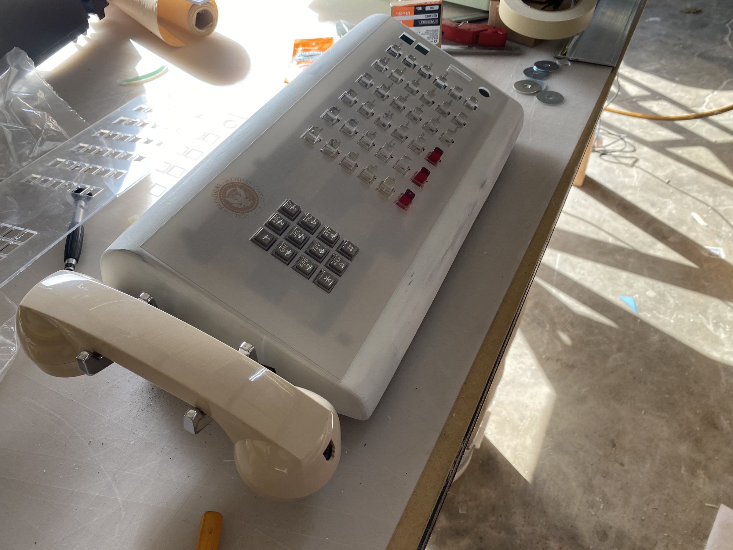 The switchboard phone mid-build on the workbench: the unpainted body with rows of clear lucite line buttons and three red ones, the cream handset cradled at the front, and the laser-cut button sheet lying beside it.
