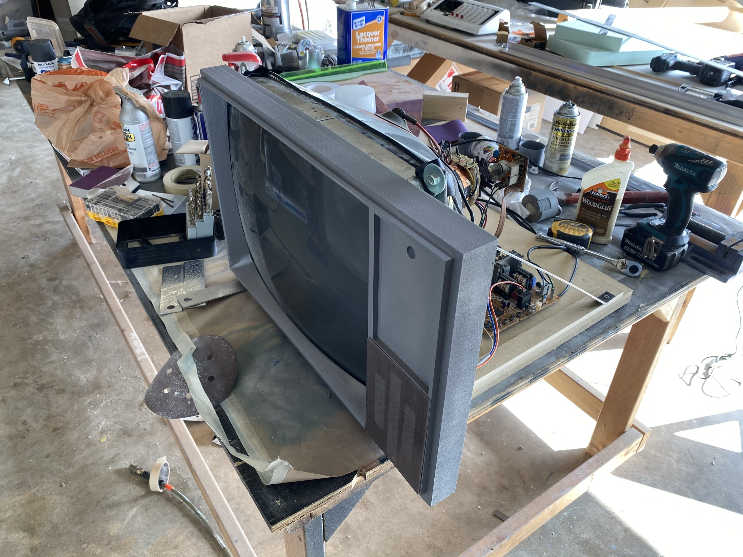 A fabricated television bezel on the workbench, its salvaged picture tube and chassis electronics exposed behind the grey CNC-cut cabinet face, among drills, glue, and spray cans.