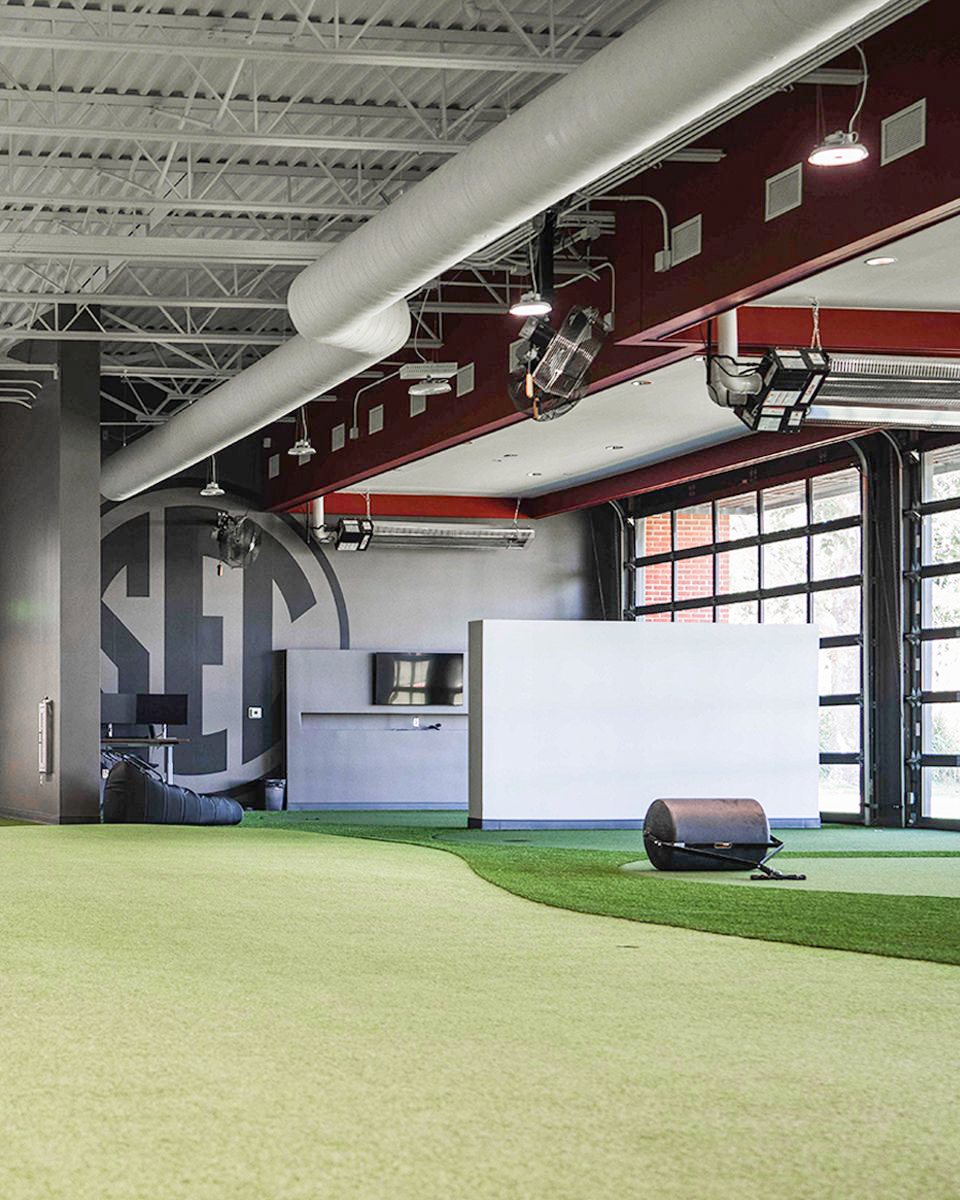 Indoor putting turf in the performance center: a tone-on-tone SEC medallion supergraphic reads faintly on the green, with glass garage doors along the wall.