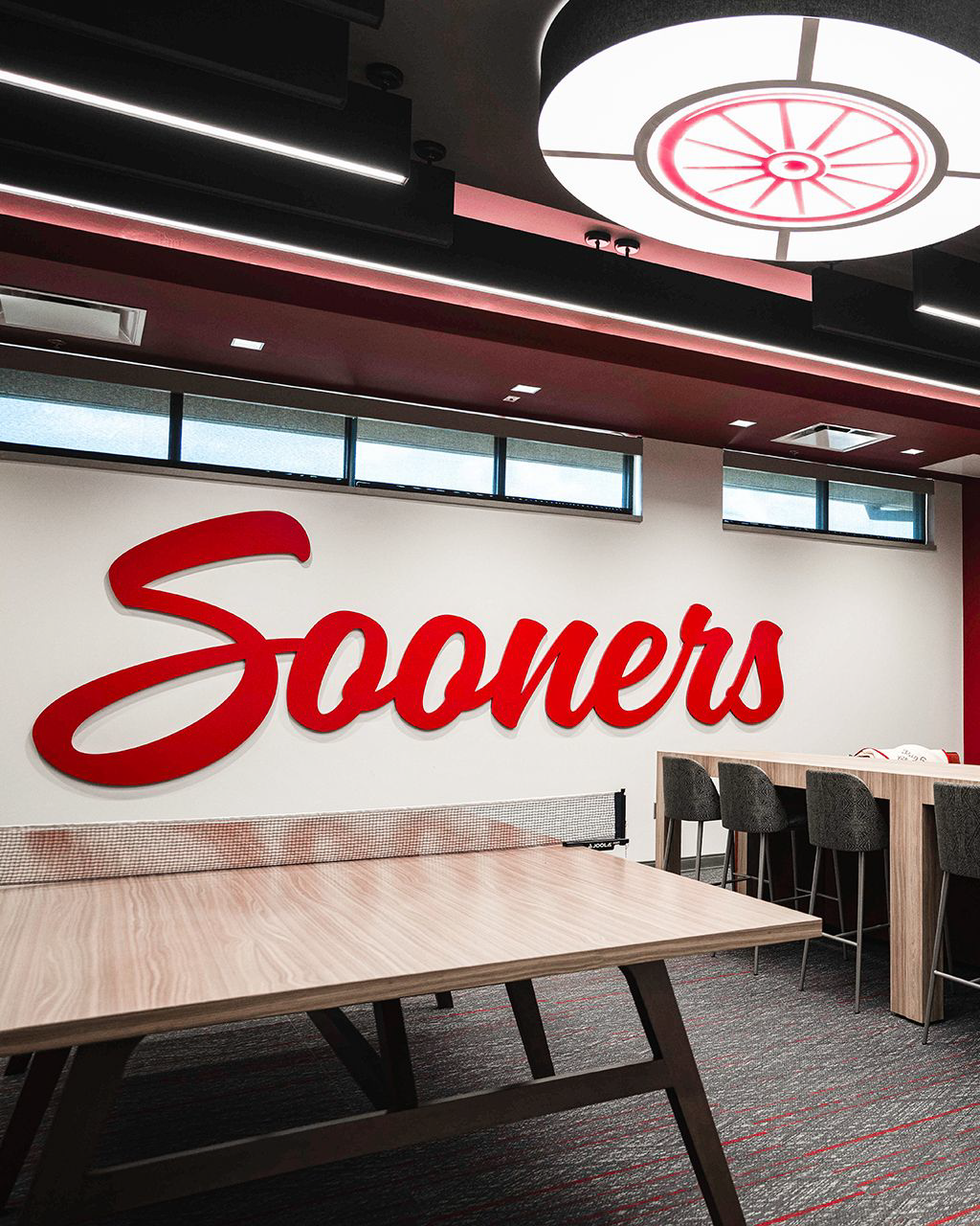Men's player lounge: a dimensional red 'Sooners' script standing off a white wall, a ping-pong table, and a circular wagon-wheel light fixture overhead.