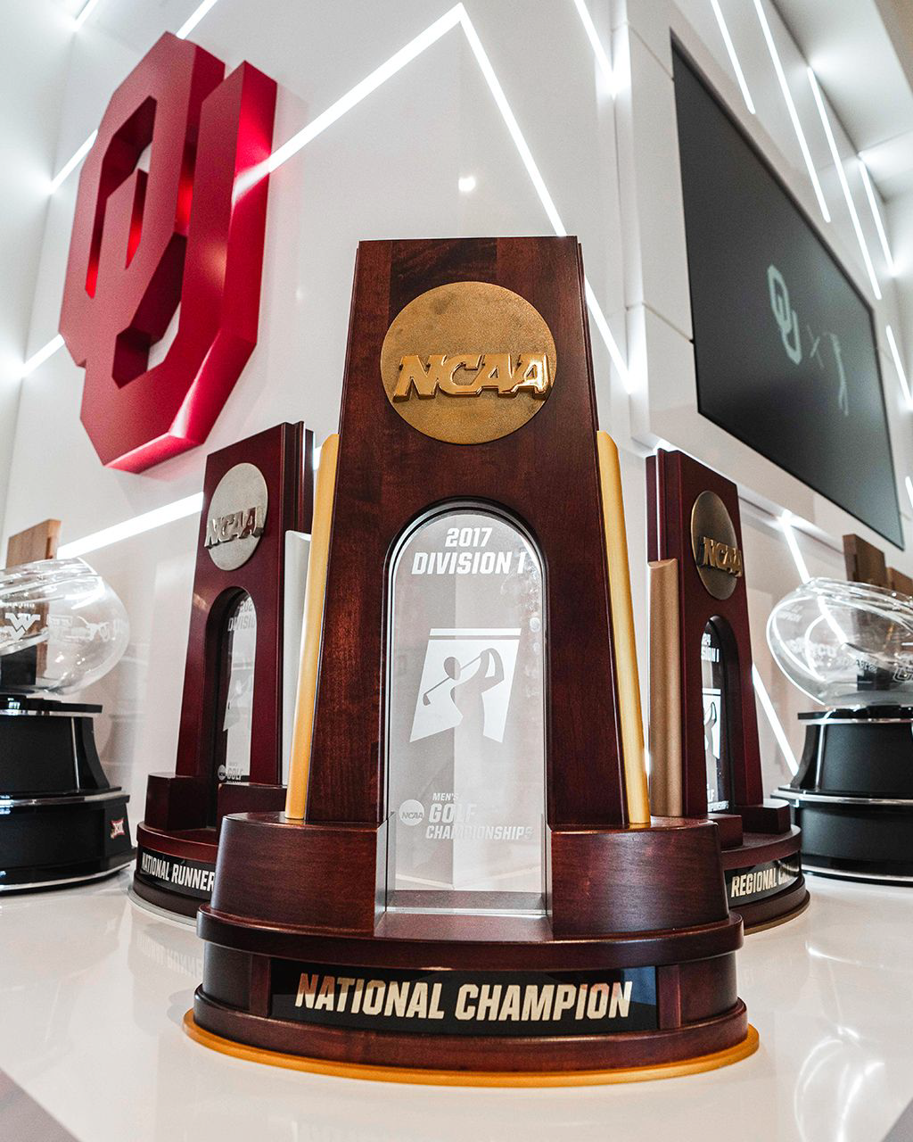 Trophy case at the nexus, close: the 2017 NCAA Division I men's golf national championship trophy flanked by NCAA and crystal-football trophies, a diagonal light-line lattice around a red OU monogram behind, and an 'OU' golfer mark on the screen above.