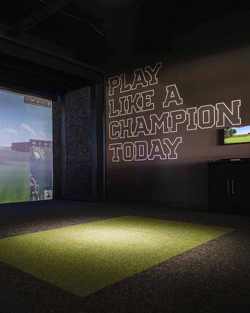 Golf simulator room, dark: a projection screen and a lit strip of turf, with 'PLAY LIKE A CHAMPION TODAY' in tone-on-tone outline block letters on the wall.