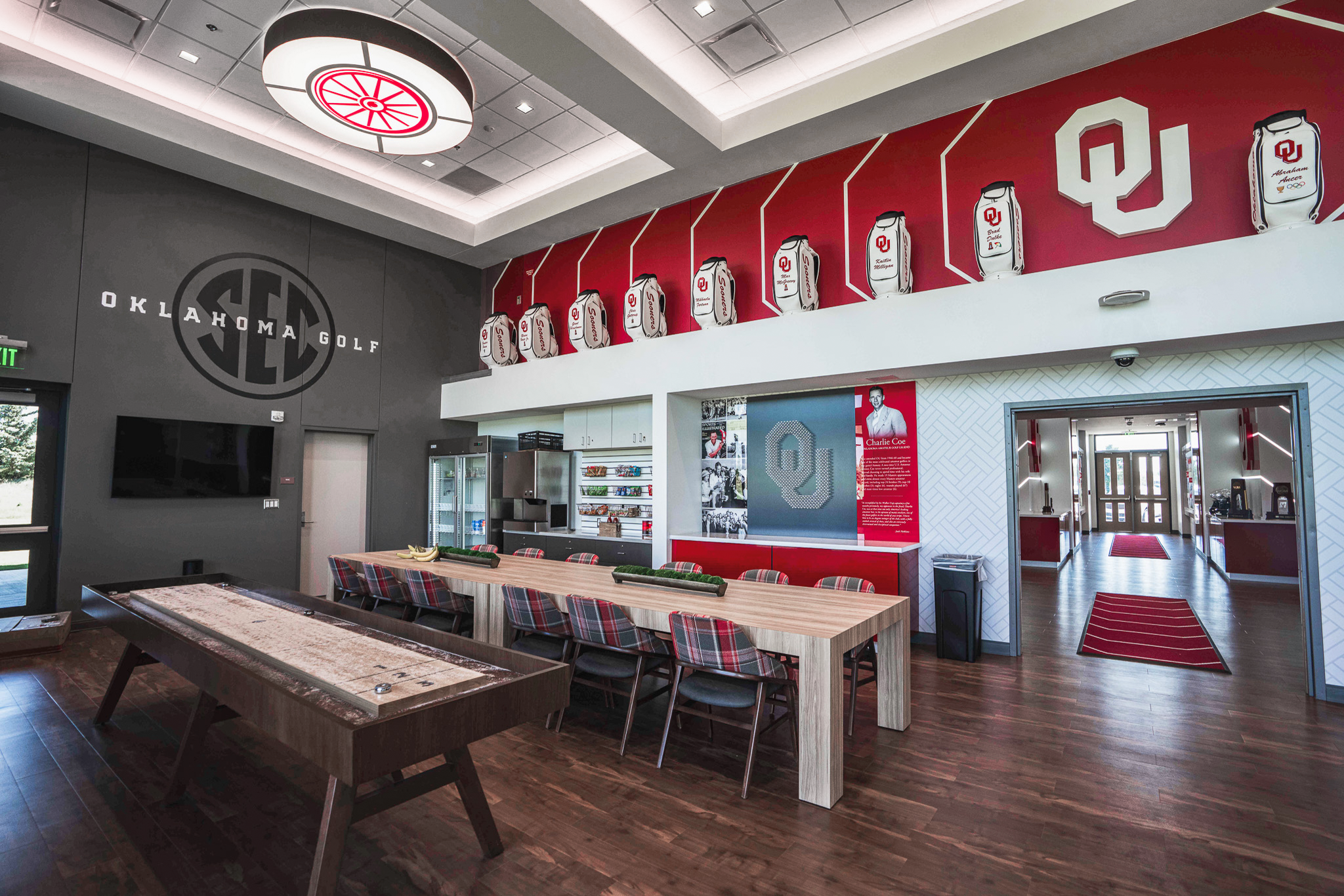 The club lounge, wide: a row of tour bags in white O-profile niches along a giant soffit under a large white OU monogram, 'OKLAHOMA GOLF' and a tonal SEC medallion on a charcoal wall, a wagon-wheel light fixture and communal table, and the Charlie Coe heritage wall at right with its golf-tee OU monogram.