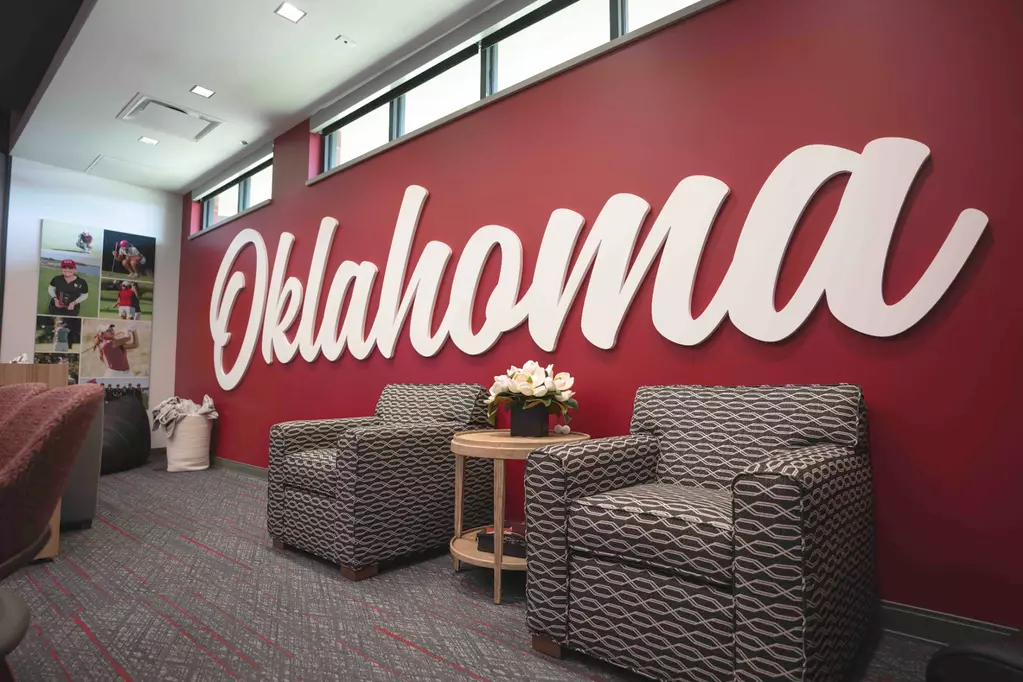 Women's lounge script wall: a white dimensional 'Oklahoma' script on red, the counterpart to the men's red 'Sooners,' with patterned armchairs and a photo-collage column.
