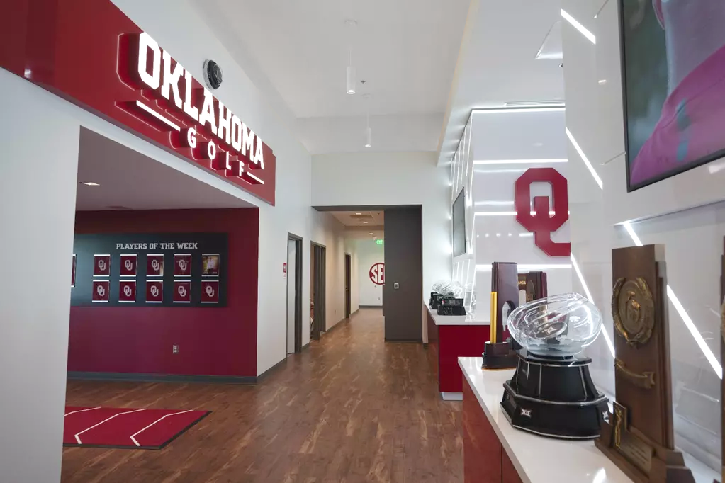 Nexus looking toward a wing: a red dimensional OKLAHOMA GOLF header with the O-profile worked into the letters, a Players of the Week display of blank OU-monogram cards beyond, an O-profile floor mat, and a tonal SEC roundel on the door at the corridor's end.