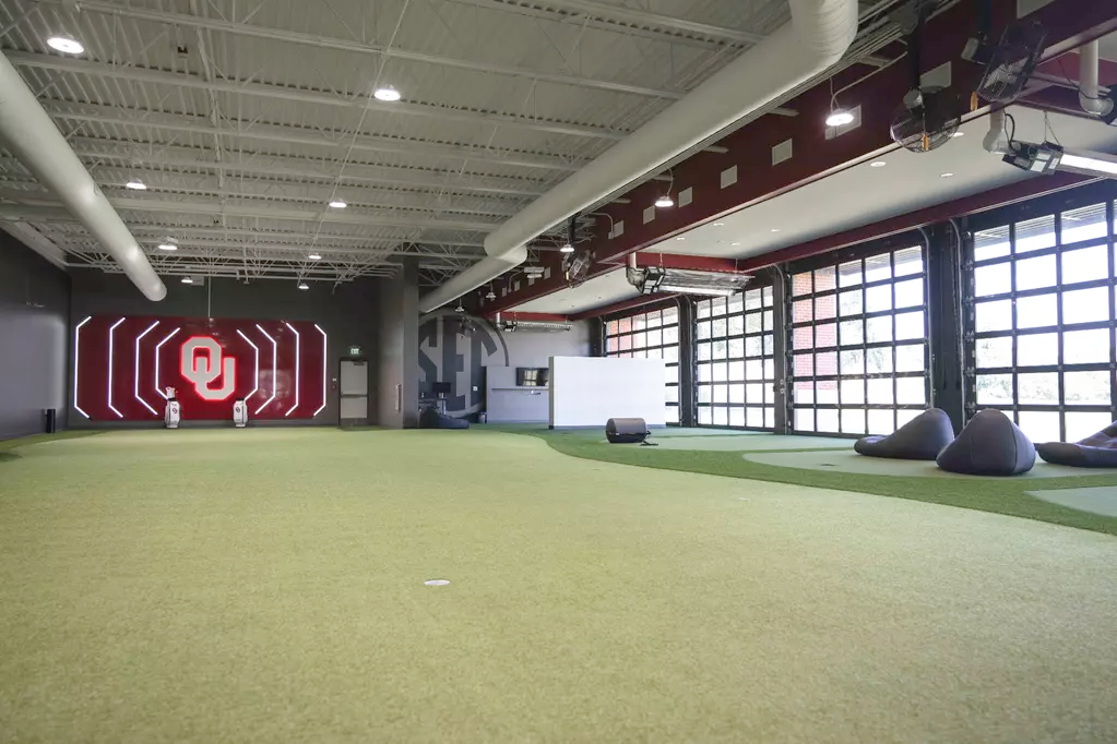 The performance center's brand wall: illuminated white O-profile outline shapes flanking a lit OU monogram on a red feature wall, staff bags beneath, a tonal SEC medallion ghosted on grey, and glass garage doors alongside.