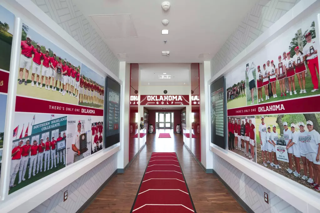 Entry corridor toward the nexus: team-photo murals on the men's and women's walls, red 'THERE'S ONLY ONE OKLAHOMA' bands, dark schedule panels, herringbone wallcovering, and a custom striped floor runner.