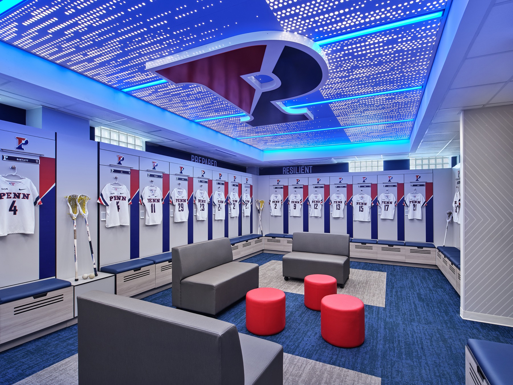A locker room under a luminous perforated ceiling carrying the red-and-blue split P, open lockers with hanging white jerseys around three walls, gray sofas and red ottomans at the center of a blue carpet.