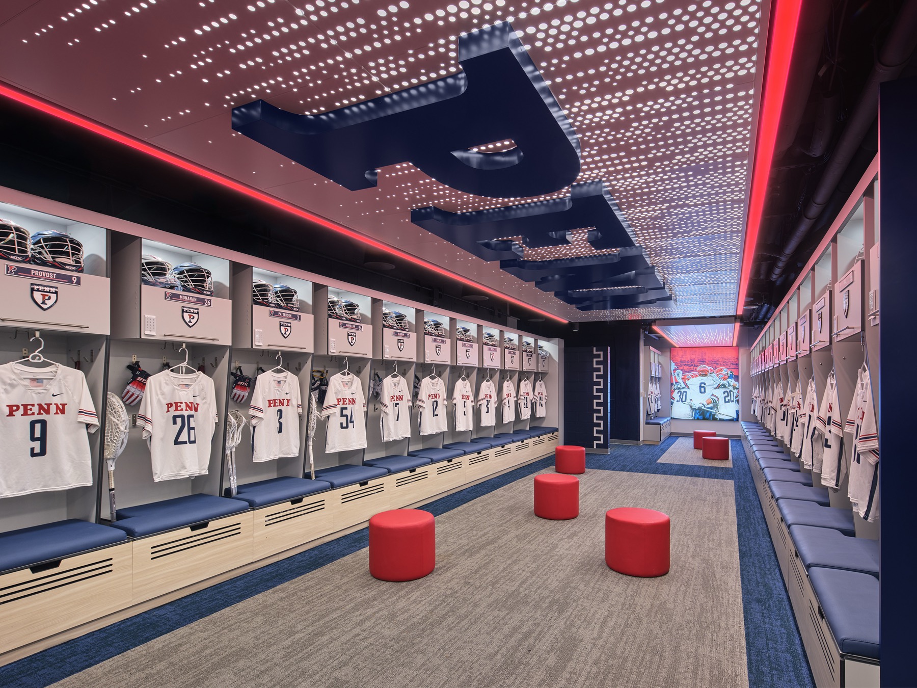 A long locker room under a perforated luminous ceiling with navy P letterforms suspended from it; helmets and gloves racked above white jerseys, benches down both sides, red ottomans on the center carpet.