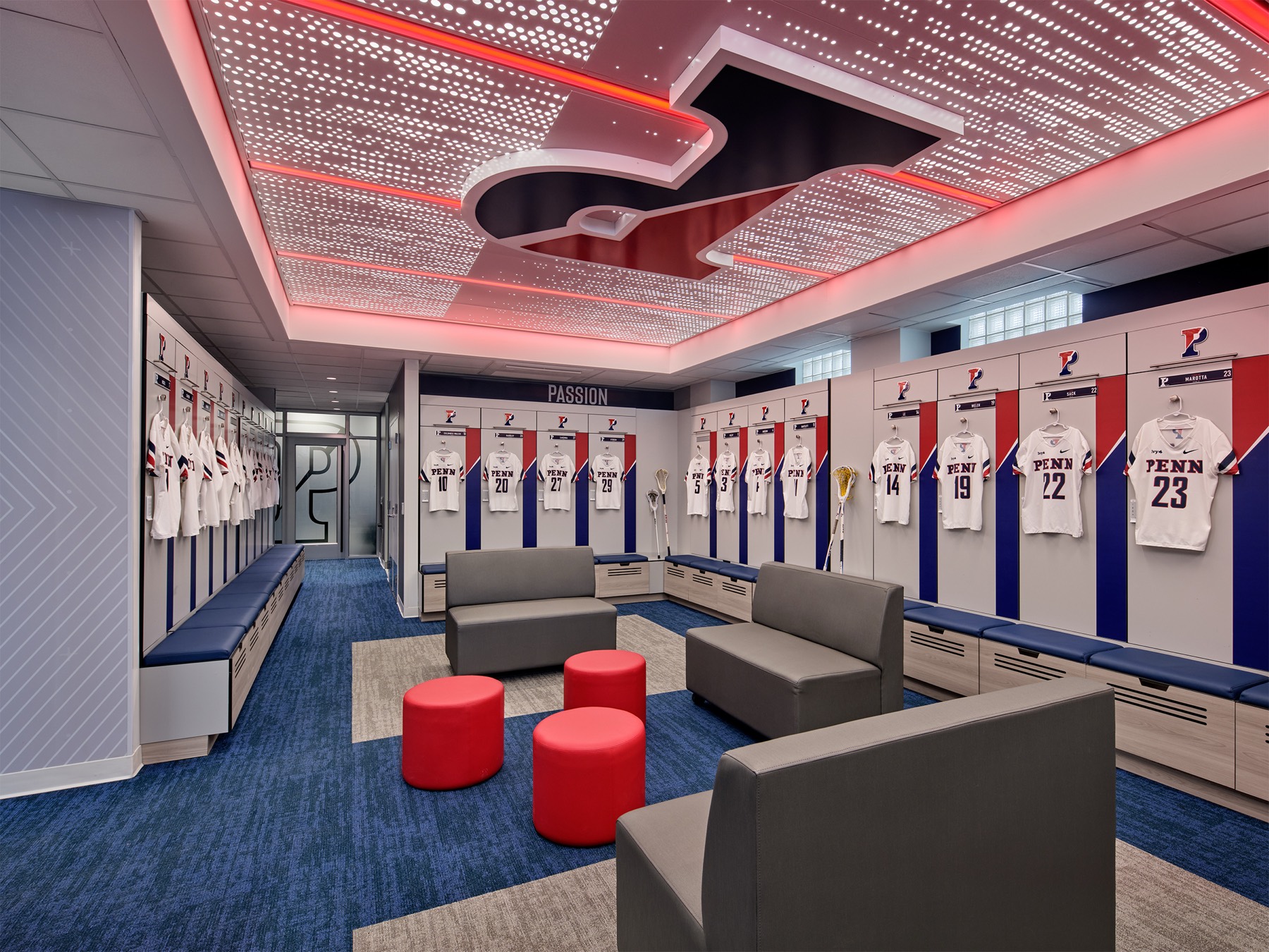 A locker room under red perimeter light: a dark letterform overhead in a field of perforated light, the word PASSION above red-white-and-blue lockers, red ottomans and gray seating on blue carpet.