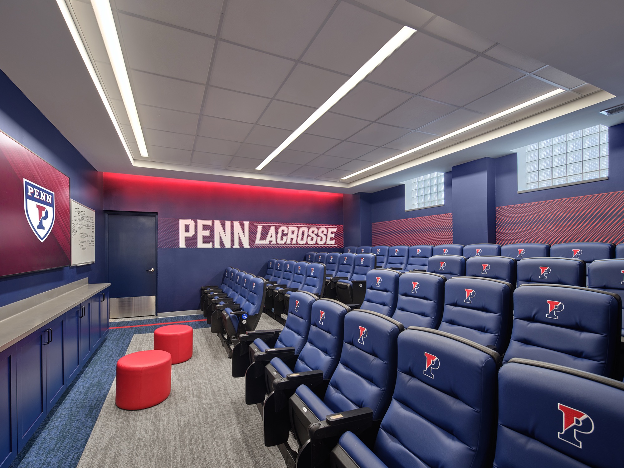 A tiered team room: rows of blue reclining seats with the P on each headrest, a red PENN LACROSSE band across the front wall, a whiteboard and monitor at the head of the room.